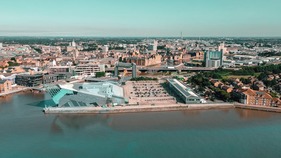 An aerial view of Kingston upon Thames showcasing a mixture of modern and historic buildings, with a prominent contemporary architectural structure located near the riverbank. The area features a parking lot with several cars, surrounded by residential and commercial buildings, some with red brick facades, and a network of roads connecting the city. The river runs along the foreground of the image, reflecting the surrounding environment. In the distance, the cityscape extends with various high-rise and mid-rise structures, under a partly cloudy sky with soft natural light. This view highlights Kingston's urban landscape, with diverse architectural styles and a vibrant riverside location, relevant to house removals and home relocation services provided by [COMPANY_NAME] in Kingston upon Thames, as seen on the page for affordable house removals KT1.