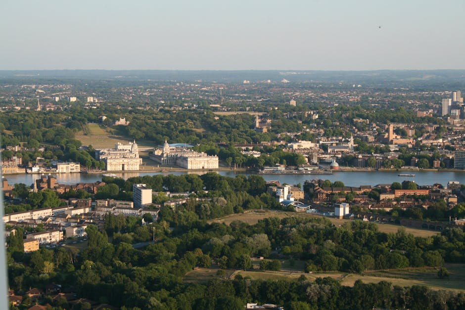 An aerial view of a densely built urban area in Kingston upon Thames, showing a mix of terraced houses, commercial buildings, and green spaces. The scene includes a waterway on the right side with small boats docked along the shoreline, adjacent to a paved walkway and a parking area. In the foreground, a busy street with moving cars, vans, and a small blue truck is visible, along with a roundabout and several intersections. To the left, there is a section of road with trees and a small grassy patch, indicating a possible site for home relocation and furniture transport activities. The image captures the typical layout of residential and commercial properties within a river-side town, highlighting the logistical complexity of house removals and packing and moving operations that a professional service like [COMPANY_NAME] would handle in Kingston upon Thames. Natural daylight and clear weather illuminate the scene, emphasizing the urban environment and transportation infrastructure involved in house removals.