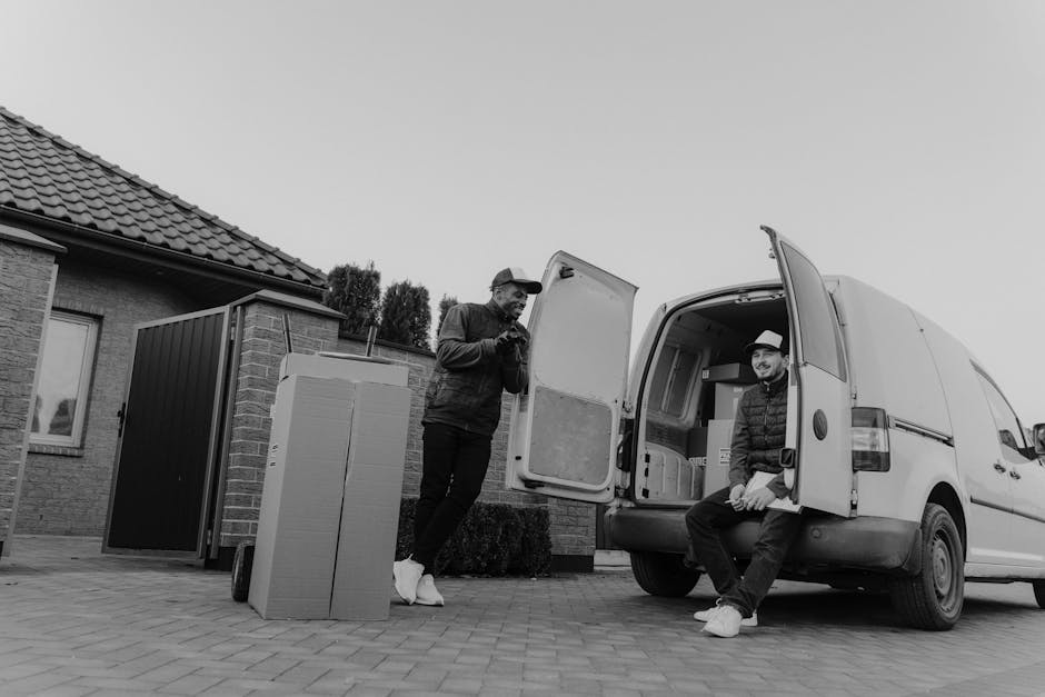 Two men are engaged in a home relocation scene outside a residential property with a brick exterior and a tiled roof. One man, dressed in dark clothing and a cap, is standing beside a large cardboard box and a plastic-wrapped item, apparently preparing to load or unload it. The second man, wearing a hat and a jacket, is sitting on the edge of an open cargo area of a white panel van, which is positioned on a paved driveway adjacent to the house. The van's rear doors are wide open, revealing interior space with packaging materials and possibly additional boxes or furniture. A small wooden shed and a modern housing structure are visible in the background. The lighting suggests daytime conditions, and the scene captures the logistical details involved in furniture transport and packing during a professional move, as performed by [COMPANY_NAME], demonstrating the loading process typical of house removals services.
