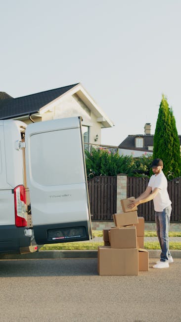 Two men are engaged in a home relocation scene outside a residential property with a brick exterior and a tiled roof. One man, dressed in dark clothing and a cap, is standing beside a large cardboard box and a plastic-wrapped item, apparently preparing to load or unload it. The second man, wearing a hat and a jacket, is sitting on the edge of an open cargo area of a white panel van, which is positioned on a paved driveway adjacent to the house. The van's rear doors are wide open, revealing interior space with packaging materials and possibly additional boxes or furniture. A small wooden shed and a modern housing structure are visible in the background. The lighting suggests daytime conditions, and the scene captures the logistical details involved in furniture transport and packing during a professional move, as performed by [COMPANY_NAME], demonstrating the loading process typical of house removals services.