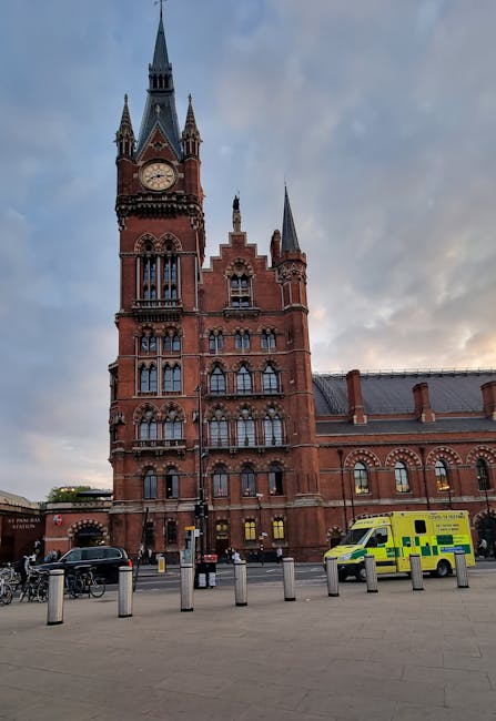 A large, historic red-brick building with Gothic architectural features, including multiple pointed spires, arched windows, and a prominent clock tower, situated in an urban setting during daylight with partly cloudy skies. In the foreground, there is a paved area with several metal bollards, a row of bicycles parked along the pavement, and a yellow and green emergency vehicle parked near the building. This scene likely depicts a well-known landmark in Kingston upon Thames, with visibility of municipal or emergency service vehicles. The image relates to house removals and transport services, showcasing the environment where furniture transport and home relocation logistics could take place, supported by [COMPANY_NAME]'s removal service.