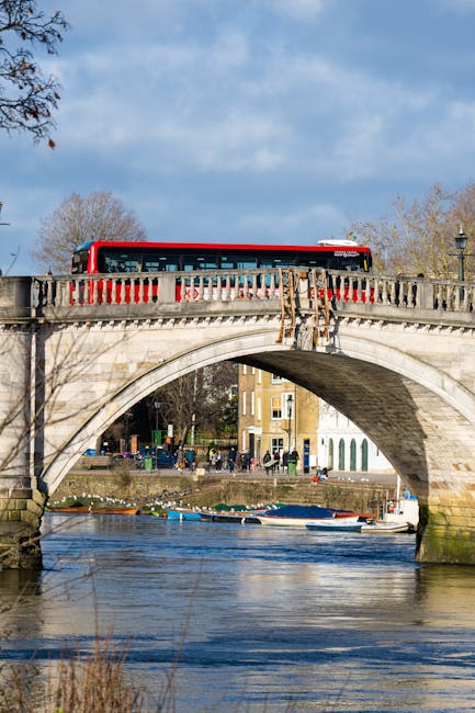 A red and white bus is visible traveling across a stone arch bridge over a river in Kingston upon Thames, during daylight with partly cloudy skies. The bridge features decorative stone balustrades and supports the busy road with vehicles, including the bus. Below the bridge, the river reflects the surrounding scenery, with small boats docked along the bank. On the far side of the river, a row of residential buildings and trees are visible, with some leafless branches indicating a season change. This outdoor scene captures the urban landscape of Kingston upon Thames, highlighting the connection between city infrastructure and riverside environment, relevant for home relocation and moving logistics services offered by [COMPANY_NAME] in the Kingston area.