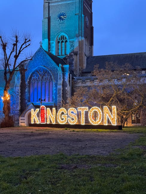 A large illuminated sign spelling 'KINGSTON' with individual letters lit in white and red lights, situated on a grassy area in front of a historic stone church with tall stained glass windows and a clock tower, during twilight or early evening. A leafless tree is visible on the left side of the church, and the scene captures the exterior of the church with a clear sky overhead. This setting is relevant to house removals and relocation services in Kingston upon Thames, where [COMPANY_NAME] may assist with the transport and handling of furniture and household items during a home relocation, set against the backdrop of a landmark feature of the area.