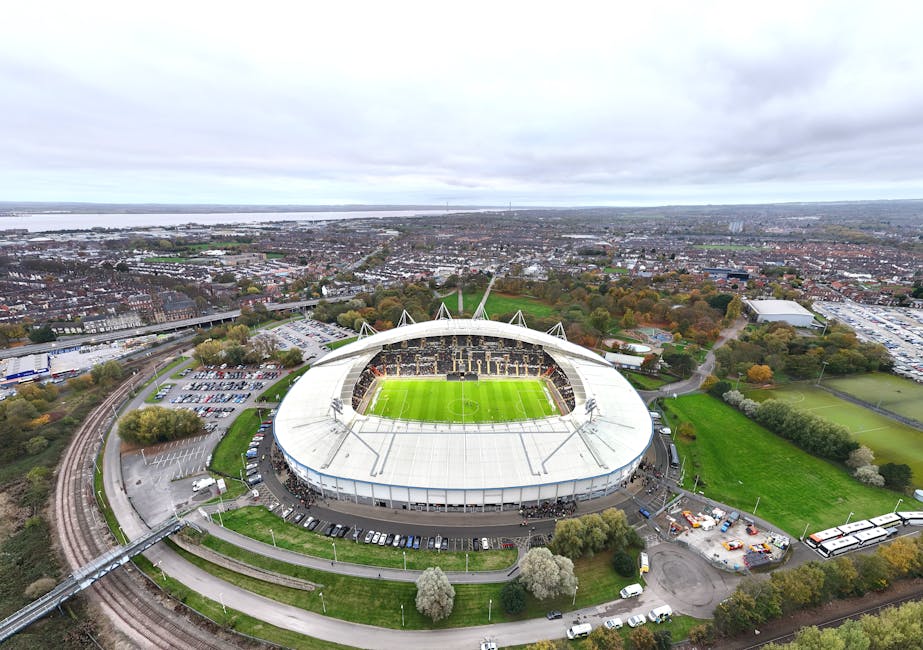 An aerial view of a large football stadium with a bright green pitch and surrounding seating areas, situated in an urban area with roads, parking lots, and parking structures nearby. The stadium has a modern design with white roofing and structural supports extending over the seating sections. Adjacent to the stadium, there are green fields and trees, with a mix of residential and commercial buildings in the background. The image showcases a wide perspective, highlighting the stadium's prominent size in relation to the surrounding cityscape, which includes multistory buildings, roadways, and a river in the distance. The overcast sky provides diffused lighting over the scene, capturing the environment in a neutral and professional manner, relevant for topics related to house removals, furniture transport, or home relocation logistics, as discussed by manandavankingstonuponthames.co.uk.