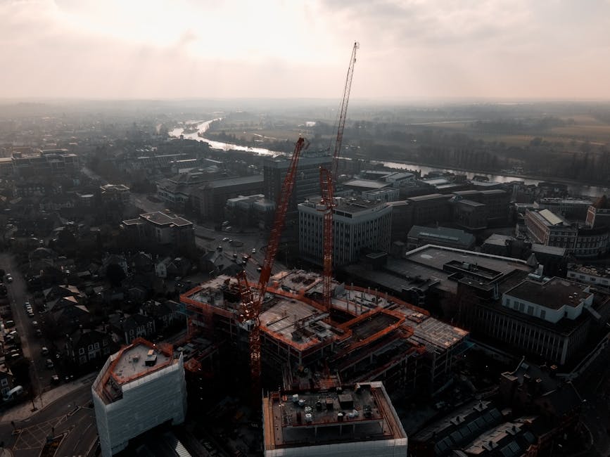 An aerial view of a multi-storey building under construction in Kingston upon Thames, with two large orange tower cranes positioned on the roof. The construction site is surrounded by surrounding urban infrastructure, including office buildings, roads, and residential areas. The scene is set during daylight with overcast skies, and a river flowing in the background beyond the cityscape. The image captures the ongoing building process, highlighting the loading and lifting activities involved in modern home relocation and furniture transport as managed by companies like manandavankingstonuponthames.co.uk, with emphasis on safe handling of construction materials and equipment.