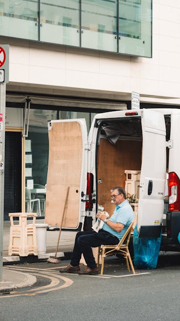A man seated on a wooden chair next to a white panel van parked on a street, engaged in reading or reviewing documents. The van's rear doors are open, revealing an interior filled with packing materials, cardboard boxes, and furniture components prepared for a home relocation. A large wooden panel, possibly a door or a piece of furniture, is leaning against the van's side. In the background, a modern building with glass windows and a storefront can be seen, along with a partially visible street sign. The scene depicts the loading process typical of furniture transport and packing as part of professional removals services provided by manandavankingstonuponthames.co.uk, reflecting the logistics involved in house removals and moving support services.