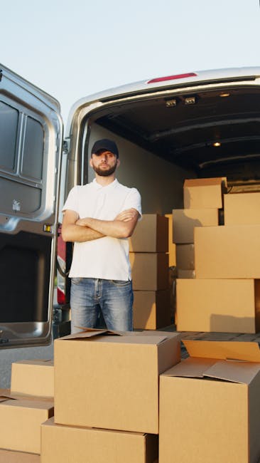 A man wearing a white polo shirt, jeans, and a black cap stands with arms crossed beside an open van loaded with multiple cardboard moving boxes of various sizes, some placed on the pavement and others within the vehicle. The van's interior is illuminated, revealing additional boxes stacked inside, awaiting transport during a home relocation or furniture transport process. The background features a clear sky, and the scene is set on a street or driveway, illustrating the packing and loading phase of a professional removals service. This image is associated with house removals and moving logistics provided by [COMPANY_NAME], emphasizing efficient packing, loading, and transportation as part of a comprehensive moving service, such as those offered through Kingston Station removals and man and van operations.