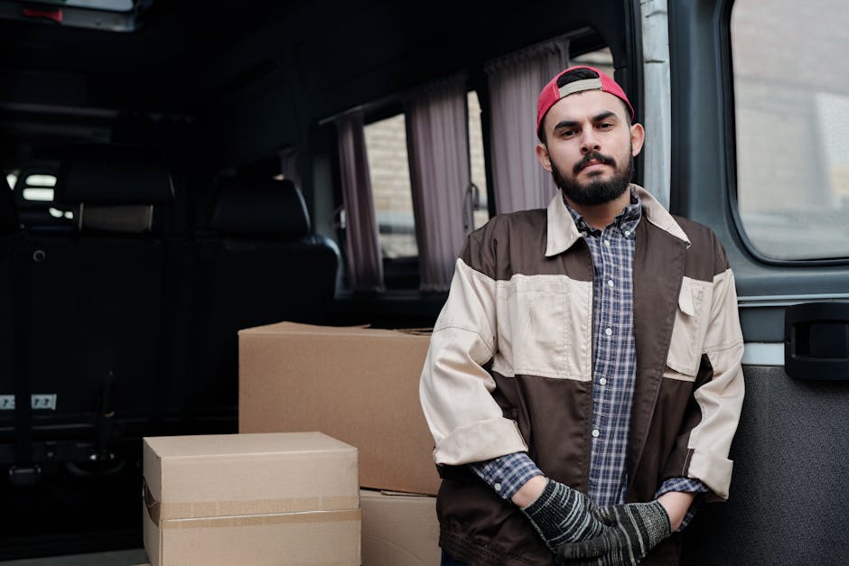 A man seated on a wooden chair next to a white panel van parked on a street, engaged in reading or reviewing documents. The van's rear doors are open, revealing an interior filled with packing materials, cardboard boxes, and furniture components prepared for a home relocation. A large wooden panel, possibly a door or a piece of furniture, is leaning against the van's side. In the background, a modern building with glass windows and a storefront can be seen, along with a partially visible street sign. The scene depicts the loading process typical of furniture transport and packing as part of professional removals services provided by manandavankingstonuponthames.co.uk, reflecting the logistics involved in house removals and moving support services.