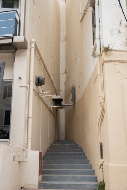 A narrow indoor staircase with red-tiled steps and white railings on both sides, leading up to an upper floor in a residential building. The staircase is situated next to a large window on the right,