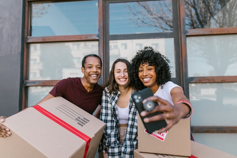 Three individuals standing outdoors in front of a modern building with large windows, engaging in a house move. The man on the left, with dark skin and short hair, is holding a cardboard box sealed with red tape, labeled 'home removals.' The woman in the middle, with light skin and long straight hair, is smiling and standing close to the other two. The woman on the right, with dark skin and curly hair, is holding a smartphone and taking a selfie while smiling broadly. She is also carrying a cardboard box. The scene suggests a cheerful moment during packing or home relocation, with moving boxes and packaging materials visible, supporting the context of house removals by [COMPANY_NAME], involved in furniture transport and packing during student moves to Kingston University for the 2026 academic year.