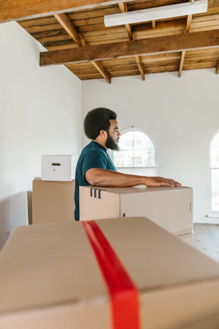 A man with dark hair and a beard, wearing a blue T-shirt, is standing inside a bright room with a wooden ceiling and exposed beams, preparing for a home relocation. He is positioned beside several cardboard moving boxes of various sizes, with some stacked on top of each other, and one box has a red packing strap across it. The room features white walls and two arched windows allowing natural light to illuminate the space. The man is holding or resting his hands on one of the larger boxes as part of the packing and furniture transport process, likely related to house removals or student move services offered by manandavankingstonuponthames.co.uk for Kingston University students undertaking term-time moves in 2026. Visible elements include the boxes, the natural light through the windows, and the wooden ceiling structure, emphasizing the packing and logistical aspects of a professional moving service.
