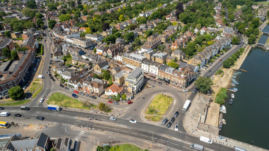 An aerial view of Kingston upon Thames showing a densely built residential area with numerous terraced houses and apartment buildings surrounded by lush green trees. In the foreground, a busy road with multiple lanes features cars, vans, and trucks moving in both directions, with some parked along the sides. Adjacent to the road, there is a small parking area with a few vehicles, and a narrow street branching off into the residential neighborhood. To the right, a riverbank with a paved walkway and small boats docked along the water's edge is visible, indicating a waterfront location. The scene is well-lit with natural daylight, highlighting the detailed urban layout and the mixture of commercial and residential buildings, which are characteristic of a typical home relocation area serviced by [COMPANY_NAME] for furniture transport and packing during house removals in Kingston upon Thames. This image illustrates the logistical environment involved in home moves, including road traffic, parcel handling, and proximity to water-based transport options for comprehensive moving services.