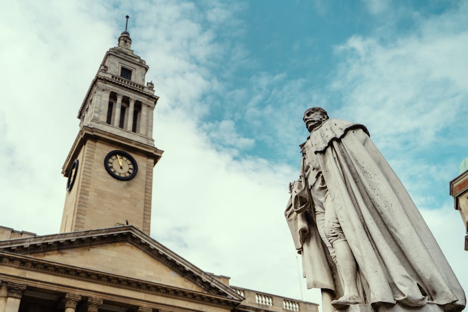 The image depicts a historic stone clock tower with a large, round clock face showing a time of approximately 3:00, situated above a classical building with decorative columns and a triangular pediment, under a partly cloudy sky. Next to the tower, a stone statue of a seated figure, wearing a long cloak and holding an object in the hand, is visible. The statue is positioned in an outdoor setting, possibly in a town square or near a public building. The scene is well-lit with natural daylight. In the context of house removals and home relocation services provided by manandavankingstonuponthames.co.uk, this image could illustrate the urban environment or iconic landmarks, emphasizing the importance of careful packing, transport, and loading in a city setting when planning a reliable move. The setting highlights the need for professional removal services to safely handle furniture, boxes, and belongings during a home relocation within or around Kingston upon Thames.